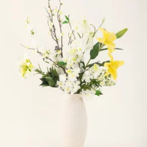 Eine Vase mit weißer Struktur zeigt die Eleganz des künstlichen Seidenblumenstraußes Sunkissed Ivory mit gelben Lilien, weißen Blüten und grünen Blättern vor einem schlichten hellen Hintergrund.