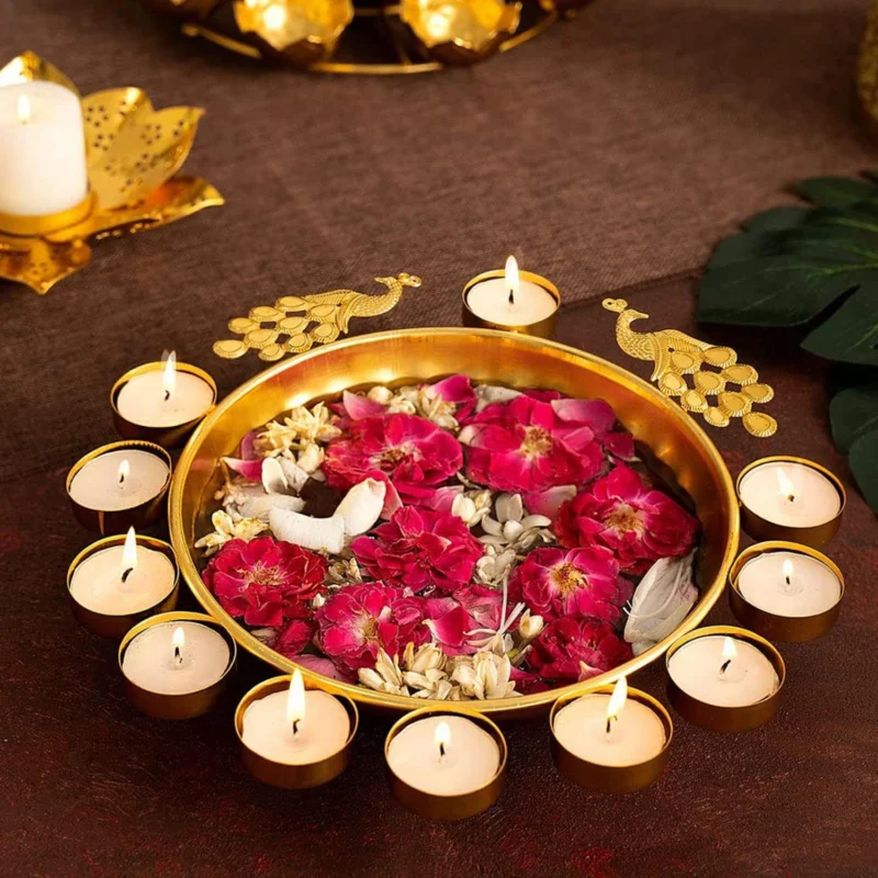 A metal urli bowl with peacock designs and tealight candle holders, adorned with pink and red flowers.