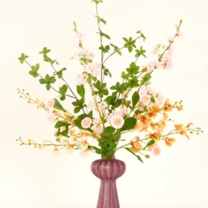 Ein Kunstblumenstrauß aus Seide von Blossom Harmony ist in einer malvenfarbenen Vase mit rosa und pfirsichfarbenen Blüten und grünen Blättern vor einem einfachen hellen Hintergrund für eine elegante Präsentation arrangiert.