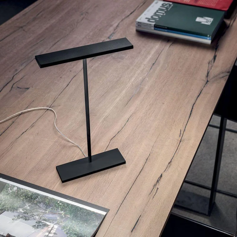 Dublight C tab LED-Tischleuchte auf einem schlichten Holztisch, umgeben von Büchern und Zeitschriften.