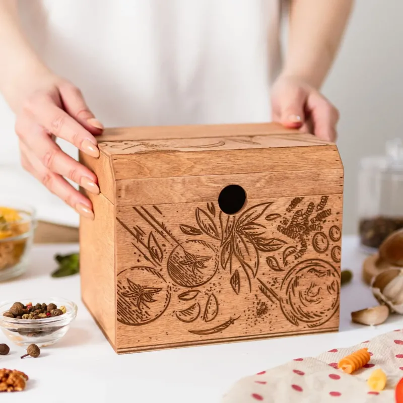 A wooden recipe box with a tomatoes design being held by a person, with recipe cards and ingredients displayed around it.