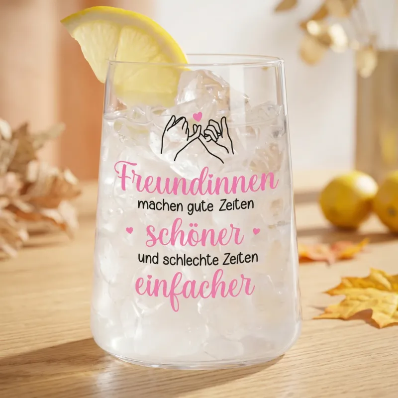 Trinkglas mit Eiswürfeln, Zitronenscheibe und Freundinnen-Motiv auf hellem Tisch