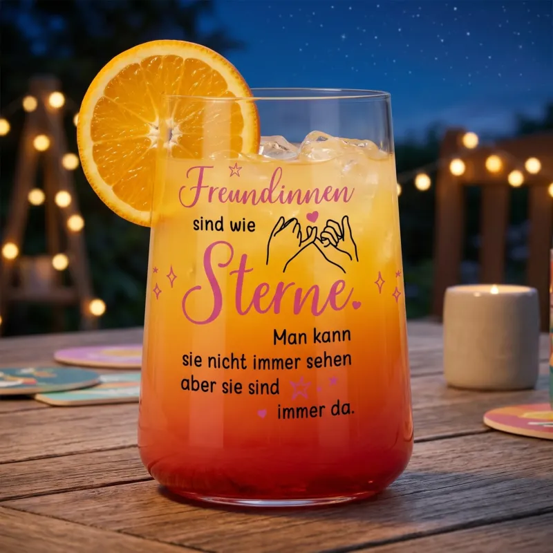 Trinkglas für Freundinnen mit rosa-schwarzem Spruch „Freundinnen sind wie Sterne“ gefüllt mit orangem Getränk und Orangenscheibe auf einem Tisch bei Abendstimmung