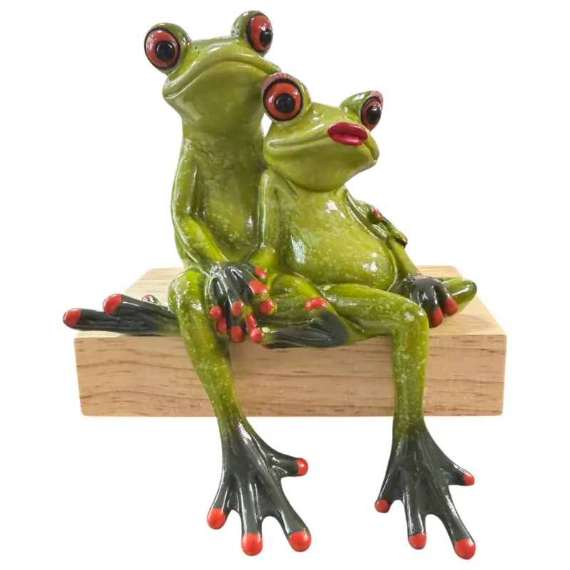 Zwei grüne Formano Frösche sitzen hintereinander auf einem hellen Holzsockel auf einem Holztisch.