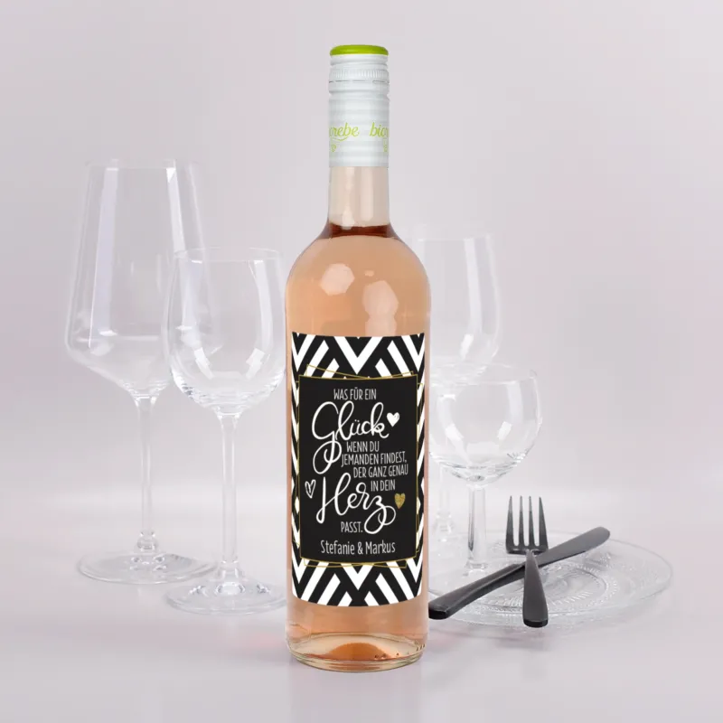 Roséweinflasche mit schwarz-weißem Etikett und goldener Schrift, Spruch „Was für ein Glück…“ und Namen Stefanie & Markus, vor Gläsern und Besteck dekoriert