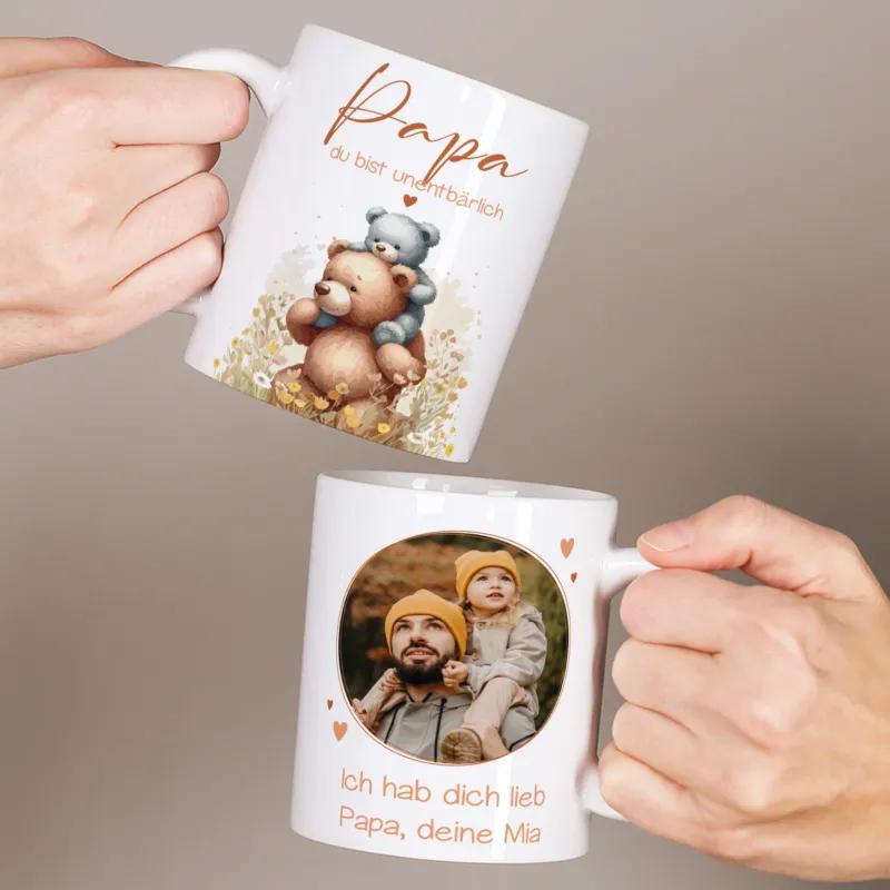 Zwei weiße Tassen werden in den Händen gehalten, links mit Bärenmotiv und dem Spruch „Papa du bist unentbärlich“, rechts mit personalisiertem Foto und Text für Papa.