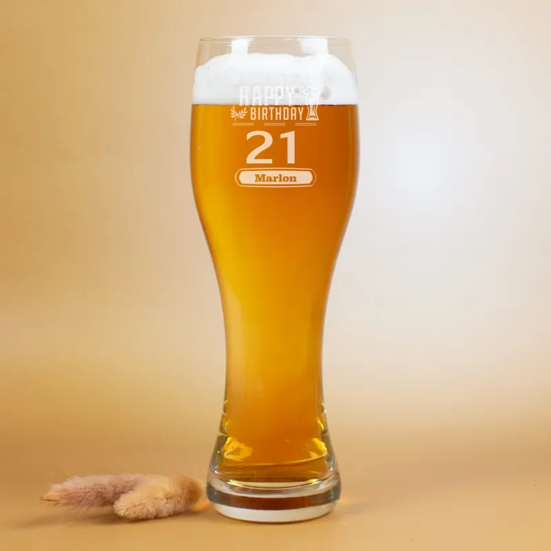 Ein gefülltes Weizenglas mit Schaumkrone, auf dem „Happy Birthday“ sowie die Zahl 21 und der Name „Marlon“ eingraviert sind. Das Glas steht vor beigem Hintergrund, daneben liegt ein kleiner Trockenblumen-Zweig.