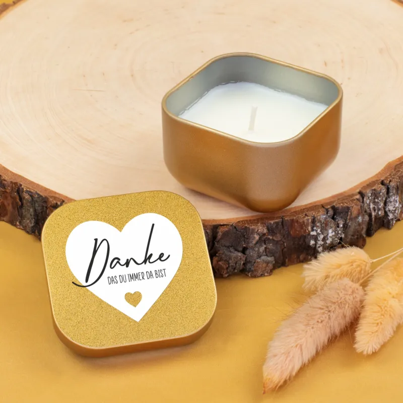 Goldene Duftkerze mit Deckel und Herzmotiv, beschriftet mit Danke das du immer da bist, vor Holzscheibe und Trockenblumen