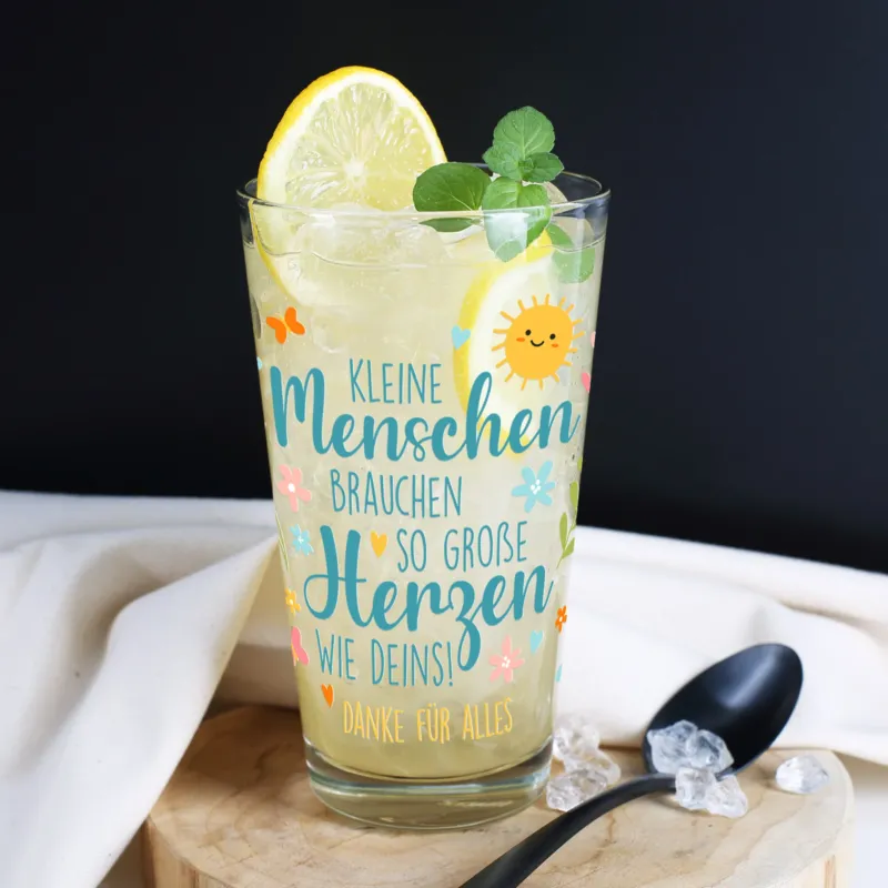 Nahaufnahme eines personalisierbaren Trinkglases mit transparenter Zitronenlimonade, dekoriert mit einer Zitronenscheibe und Minzblättern. Das Glas steht auf einem runden Holzbrett vor dunklem Hintergrund. Bedruckt mit dem Spruch „Kleine Menschen brauchen so große Herzen wie deins!“ sowie dem Zusatz „Danke für alles“. Das farbenfrohe Motiv zeigt eine lachende Sonne, Blumen, Herzen, Schmetterlinge und Blätter.