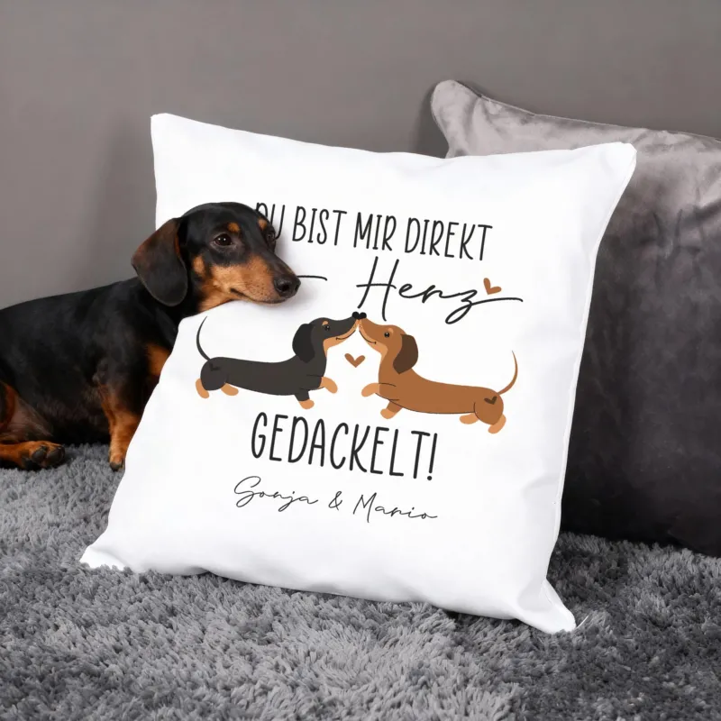 Personalisiertes Kissen mit zwei Dackeln und Herzchen, Spruch „Ins Herz gedackelt!“, Hund liegt daneben auf Sofa