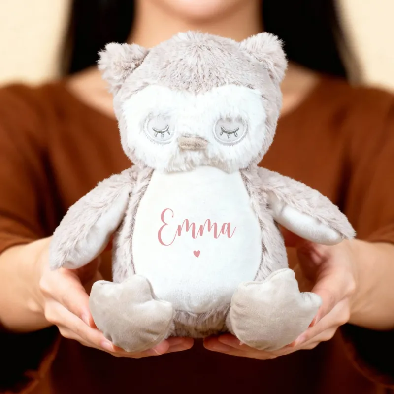 Eine Person hält eine flauschige Eule mit rosa Aufschrift „Emma“ und Herzmotiv in beiden Händen vor neutralem Hintergrund.