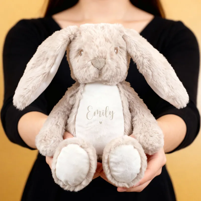 Eine Person hält einen flauschigen beigefarbenen Hasen mit der Aufschrift „Emily“ und kleinem beigem Herzmotiv auf weißem Bauch. Das Kuscheltier hat lange Ohren und einen liebevollen Gesichtsausdruck.