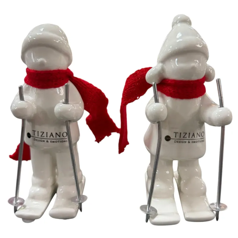 Zwei cremeweiße Tiziano Keramik-Winterkinder mit roten Schals und silberfarbenen Skistöcken vor weißem Hintergrund.